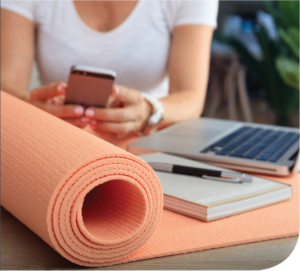 A laptop and notepad sit on top of a peach colored yoga mat, while a woman checks her phone in the background.