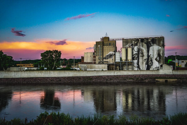 Silos with murals painted on them stand with a river in the foreground reflecting a blue and peach sunset