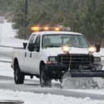 A pickup with a yellow light bar and a plow attachment clears a road of snow, with pine trees in the background.