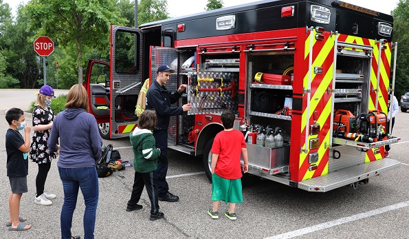 Children in summer play clothes and a woman in jeans watch as a Richfield firefighter explains equipment on a fire truck. 