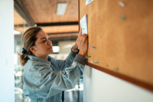 A professional woman with brown hair posts a generic notice on a cork bulletin board.