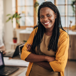 Young black businesswoman standing beside a desk in a sunny office with houseplants.