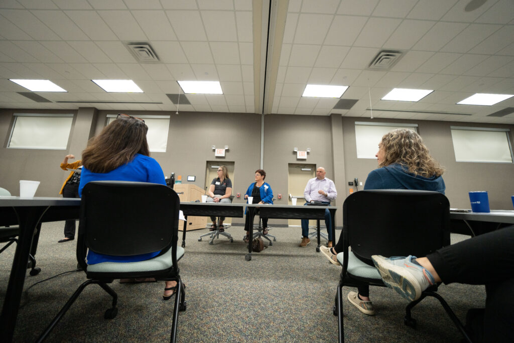 Three experienced clerk sit behind a table as the address a conference room full of city clerks.