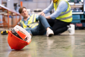 Selective focus on hard hat. Male worker appears to be injured in background, while co-worker provides help. 