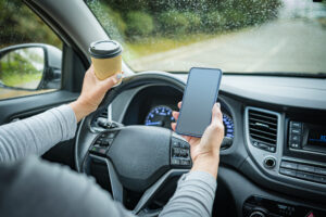 Woman holding disposable coffee cup and mobile phone while driving on wet road on a rainy day.
