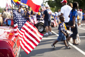 American flag on back of vehicle. The people in the background are preparing floats and rushing to join their group for the upcoming 4th of July parade.