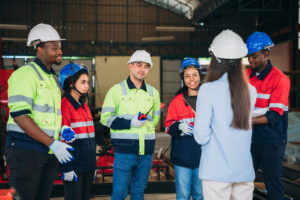 Group of workers wearing safety helmets and reflective jackets listening to a woman in a light blue blazer.