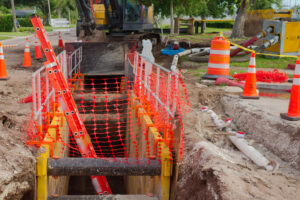 Construction site featuring safety cones, protective mesh fencing around a trench, and pipeline equipment, highlighting precaution and progress in urban infrastructure development.